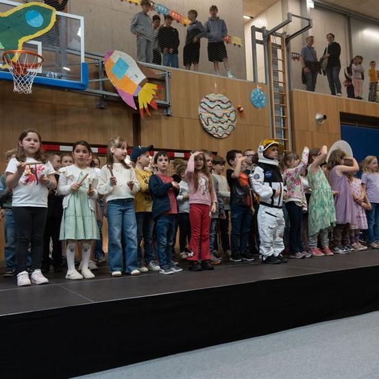 Eine Gruppe von Kindern steht auf einer Bühne, einige Erwachsene im Hintergrund und Dekorationen hängen an den Wänden.
