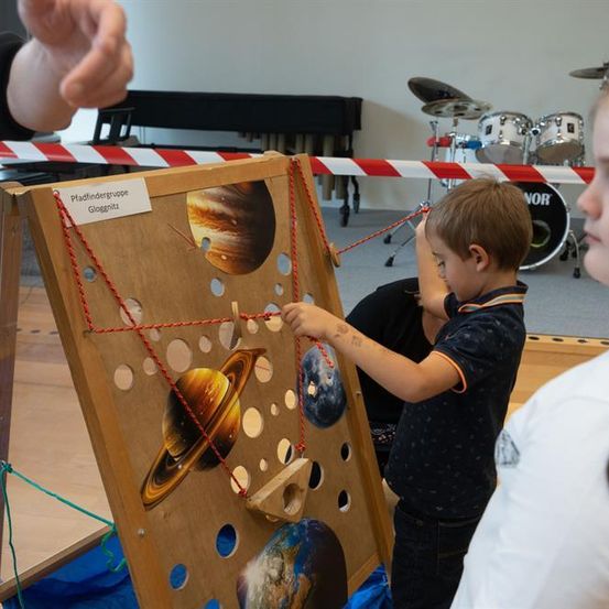 Ein Junge spielt mit einem hölzernen Lehrbrett mit verschiedenen Planeten darauf. Ein Mädchen und eine Person beobachten ihn. Hinter ihnen steht ein Schlagzeug mit einem roten und weißen Warnband.