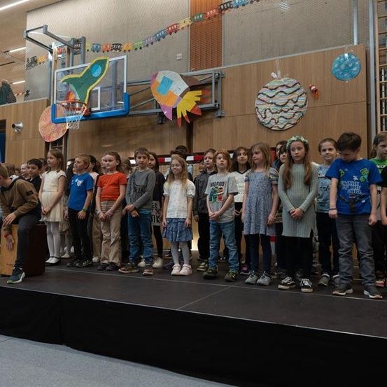 Eine Gruppe von Kindern steht auf einer Bühne, mit mehreren Dekorationen im Hintergrund, einschließlich eines Basketballkorbs.