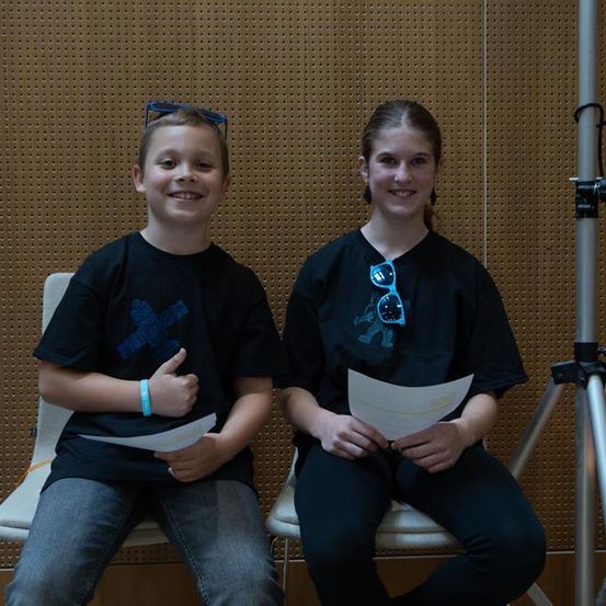 Zwei Kinder sitzen mit Papieren auf Stühlen und lächeln für die Kamera. Ein Junge gibt Daumen hoch. Ein Stativ steht hinter ihnen.