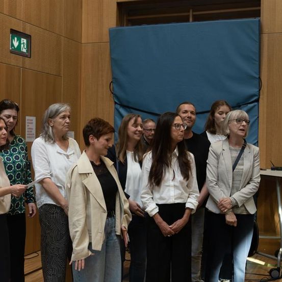 Eine Gruppe von Frauen und Männern in verschiedenen Kleidungsstücken steht zusammen in einem Raum, möglicherweise für ein Foto. Der Raum hat Holzwände und einen blauen Hintergrund.
