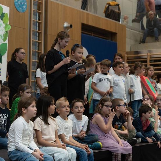 Eine Gruppe von Kindern sitzt auf der Bühne und hört aufmerksam einer Mädchen zu, die ein Mikrofon und ein Blatt Papier in der Hand hält. Andere Kinder sitzen vor ihnen, und ein Poster mit verschiedenen Zeichnungen ist an der Wand sichtbar.