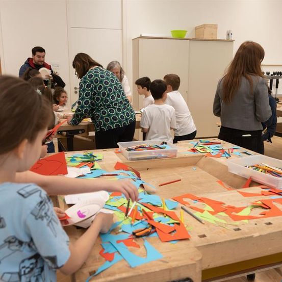 Eine Gruppe von Kindern bastelt mit farbigem Papier, Scheren und Kleber. Sie sitzen um einen Tisch herum und arbeiten an verschiedenen Kunstprojekten.