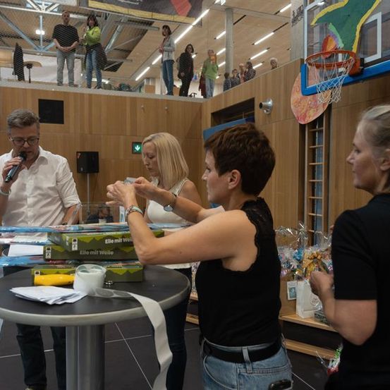 Ein Mann spricht in ein Mikrofon, während zwei Frauen ein Band und eine Box halten. Andere stehen in der Nähe, und Zuschauer sind auf einer Galerie.