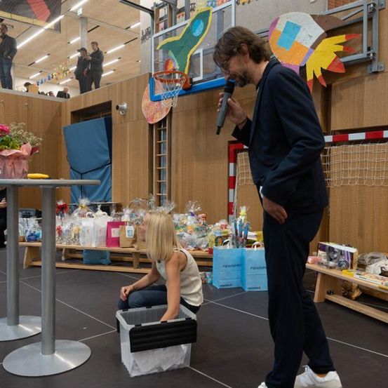 Ein Mann in einem Anzug spricht in ein Mikrofon, während ein junges Mädchen auf dem Boden sitzt und Gegenstände in einer Aufbewahrungsbox betrachtet. Hinter ihnen ist ein Basketballkorb an der Wand montiert.