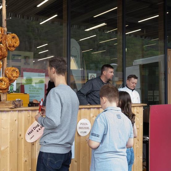 Eine Gruppe von Menschen steht vor einem Essensstand. Ein Mann hält ein rotes Schild für Hot Dogs. Ein Junge und ein Mädchen stehen vor einem Pizzaschild. Dahinter schaut ein Mann in einem weißen Hemd sie an.