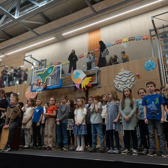 Eine Gruppe von Kindern steht auf der Bühne in einer Turnhalle, während Erwachsene von oben beobachten. Ein Basketballkorb und bunte Dekorationen sind vorhanden.