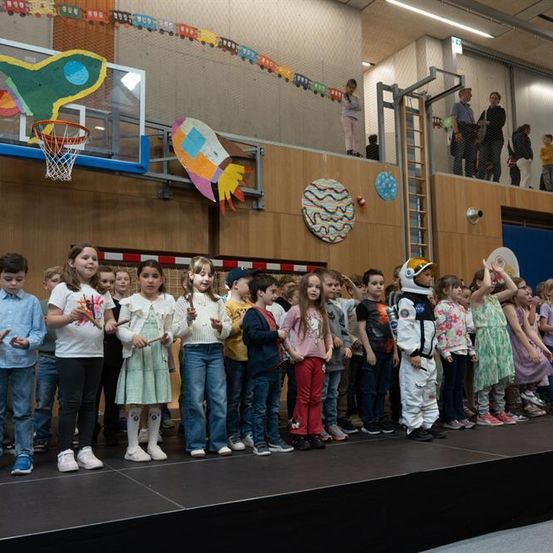 Kinder auf einer Bühne mit Weltraum- und Zugdekorationen, eines in einem Raumanzug, andere halten Musikinstrumente.