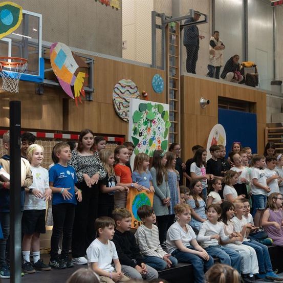 Kinder stehen auf einer Bühne in einer Turnhalle, einige halten Schilder. Hinter ihnen sind Poster und ein Basketballkorb zu sehen. Erwachsene beobachten vom Balkon aus.