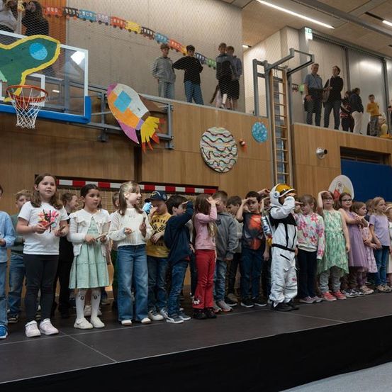 Eine Gruppe von Kindern und Erwachsenen steht auf einer Bühne mit einer Person in einem Raumanzug. Einige Kinder halten Musikinstrumente. Im Hintergrund befindet sich ein Basketballkorb, eine Rakete und eine dekorative Sonne. Leute stehen auf dem Balkon.