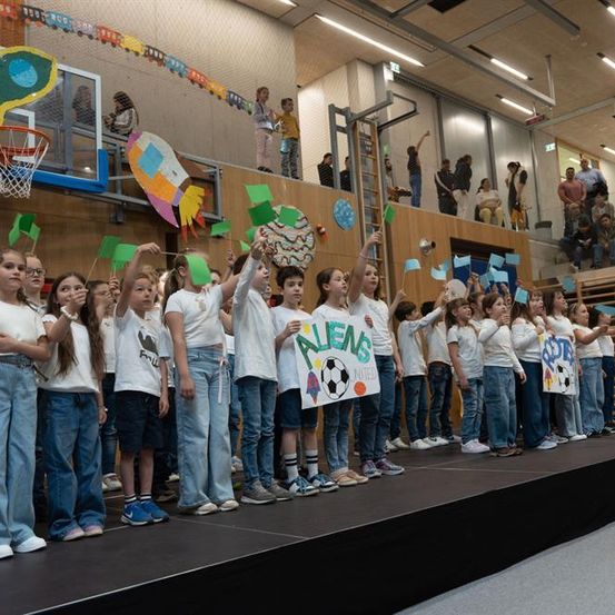 Kinder auf der Bühne halten Schilder, eines steht 'Bluens braucht', andere halten Flaggen, mit Zuschauern in einer Turnhalle.