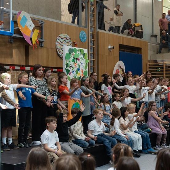 Eine Gruppe von Kindern führt auf der Bühne vor und einige klatschen, während ein Publikum auf den Tribünen zuschaut. Dekorationen und ein Basketballkorb sind im Hintergrund zu sehen.