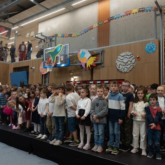 Eine Gruppe von Kindern steht in einer Reihe auf der Bühne einer Turnhalle, während Erwachsene von der Galerie zuschauen. Die Wände sind mit bunter Kunst dekoriert.