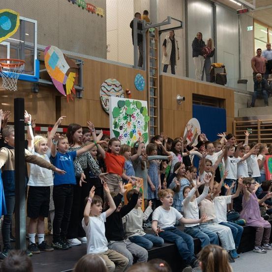 Eine Gruppe von Kindern tritt in einer Turnhalle auf. Einige heben die Hände, während andere sitzen und zusehen. Auf der linken Seite ist ein Basketballkorb zu sehen.