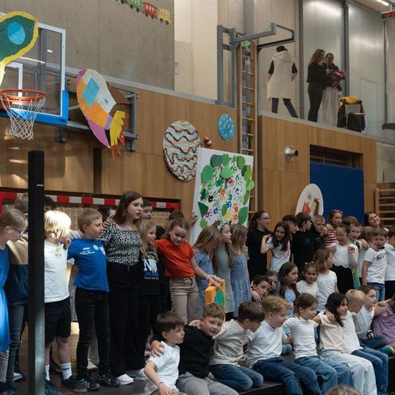 Eine Gruppe von Kindern und einer Frau steht in einem großen Innenraum. Sie sind in Reihen angeordnet, einige sitzen und andere stehen. Die Wände sind mit Kunst und Bastelarbeiten dekoriert. Ein Basketballkorb ist an der Wand montiert. Einige Leute stehen auf der oberen Ebene.
