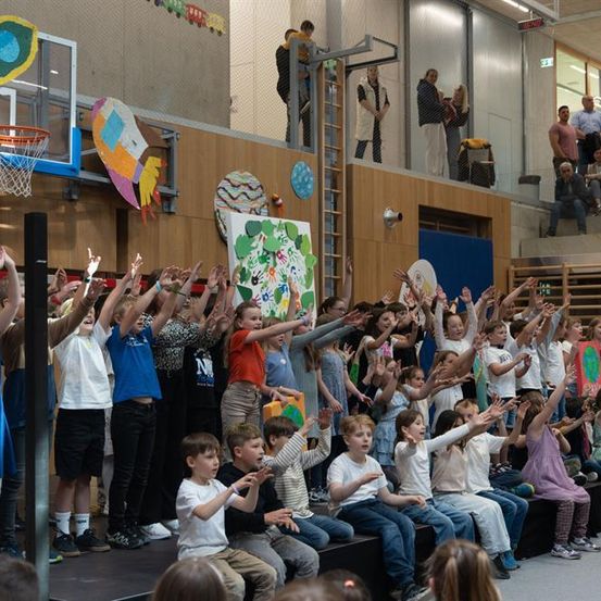 Eine Gruppe von Kindern auf der Bühne, die ihre Hände heben, verschiedene farbige Hemden und Hosen tragen. Einige sitzen auf Bänken, während andere stehen. Bunte Kunstwerke sind an der Wand hinter ihnen.