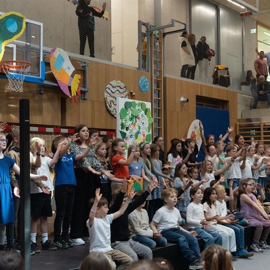 Eine Gruppe von Kindern steht auf einer Bühne und klatscht in die Hände. Einige heben ihre Hände. Sie tragen Freizeitkleidung. Dahinter stehen einige Menschen auf dem Balkon und beobachten die Kinder. Die Wand hat einen Basketballkorb, ein Poster und andere Dekorationen. Die Decke hat Lampen und eine Uhr.