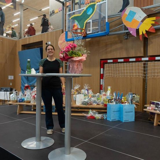 Eine Frau steht lächelnd auf einer Bühne mit zwei Tischen, umgeben von verschiedenen Gegenständen wie Blumen, Geschenken und einem Basketballkorb im Hintergrund.