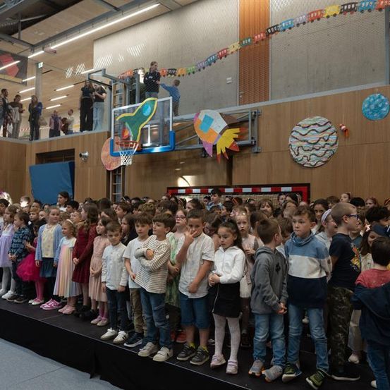 Eine große Gruppe von Kindern steht auf einer Bühne in einer Schulturnhalle. Einige tragen Turnschuhe, andere Stiefel. Ein Basketballkorb und Dekorationen schmücken den Hintergrund. Erwachsene beobachten von oben.