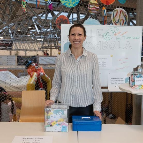 Eine Frau steht lächelnd hinter einem Tisch mit einer Box und einem blauen Kasten. Hinter ihr steht ein weißes Schild mit den Worten 'LE MBOLA'.
