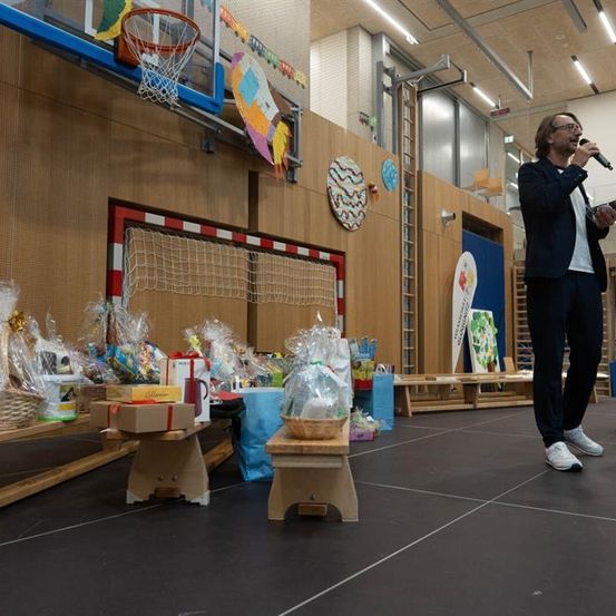 Ein Mann steht auf einer Bühne mit einem Mikrofon und hält wahrscheinlich eine Rede. Verschiedene Geschenkkörbe stehen auf dem Boden und eine Basketballkorb ist an der Wand montiert.