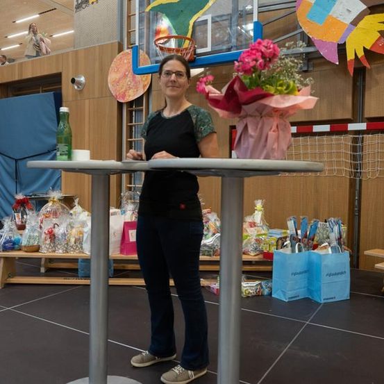 Eine Frau steht vor einem Tisch mit einem Blumenstrauß, trägt eine Brille und Turnschuhe. Hinter ihr befindet sich ein Basketballkorb, und links davon ein Regal mit verschiedenen Gegenständen. Rechts sind Taschen auf dem Boden.
