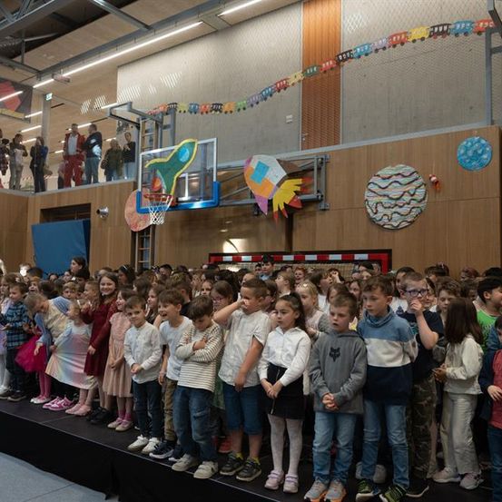 Eine große Gruppe von Kindern steht auf der Bühne in einer Turnhalle. Sie stehen in Reihen. Ein Basketballkorb ist an der Wand über ihnen.