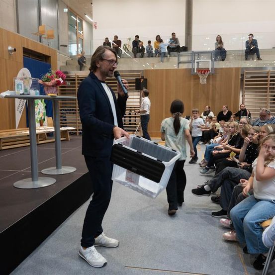 Ein Mann im Anzug hält ein Mikrofon und spricht zu einem sitzenden Publikum in einer Indoor-Sportanlage. Eine Frau in der Nähe trägt eine Aufbewahrungsbox.