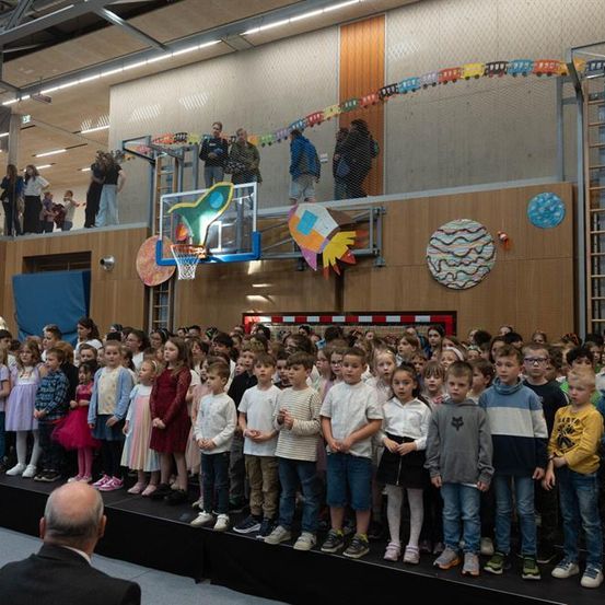 Eine Gruppe von Kindern steht auf einer Bühne, mit Erwachsenen, die von oben zusehen, und bunten Dekorationen im Hintergrund.