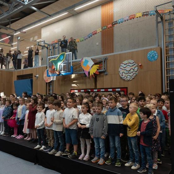 Eine große Gruppe von Kindern steht auf einer Bühne mit einem Basketballkorb und Dekorationen im Hintergrund. Einige Erwachsene beobachten von oben.