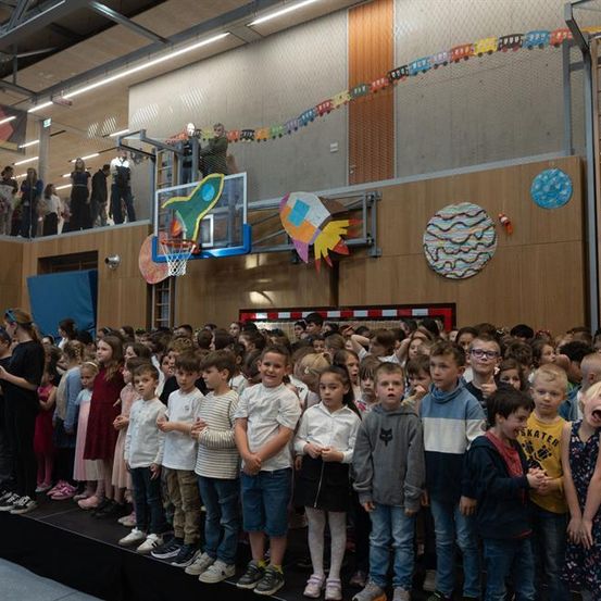 Eine große Gruppe von Kindern und Erwachsenen ist drinnen versammelt. Kinder stehen in Reihen auf einer Bühne, während Erwachsene auf einem Balkon stehen. Dekorationen und ein Basketballkorb sind sichtbar.