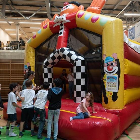 Mehrere Kinder spielen um einen bunten Clown-Aufblasbaren herum, während ein Mädchen auf der Kante sitzt. Dahinter sitzen Leute an einem Tisch auf einem Balkon.
