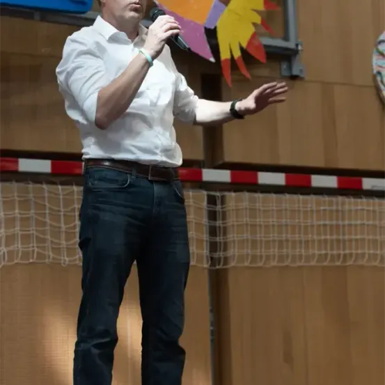 Ein Mann mit Brille, weißem Hemd und Jeans hält ein Mikrofon in einer Sporthalle. Hinter ihm ist ein Basketballkorb und ein bunter Papierdrachen zu sehen.