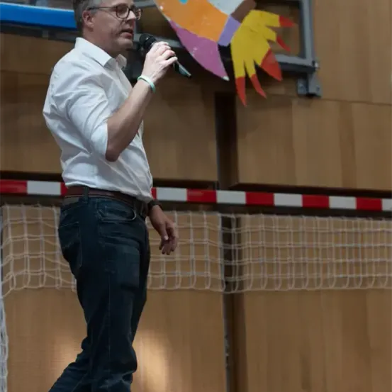 Ein Mann mit Brille und weißem Hemd steht auf der Bühne und hält ein Mikrofon. Hinter ihm ist ein buntes Raketenbild an der Wand befestigt.