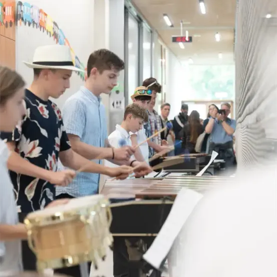 Eine Gruppe junger Jungen und Mädchen spielt Xylophon in einem Flur, während andere zuschauen. Einige Leute machen Fotos, und der Flur hat eine Wand mit hängenden Dekorationen.