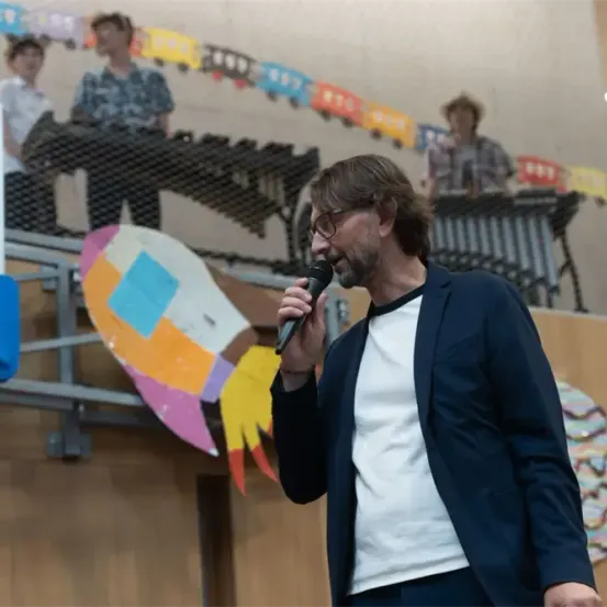 Ein Mann spricht in ein Mikrofon auf einer Bühne, mit einem Raketendesign hinter ihm und drei Musikern, die Xylophon spielen.