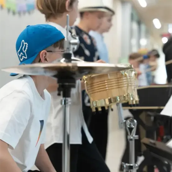 Eine Gruppe von Kindern spielt Musikinstrumente in einem Klassenzimmer. Ein Junge in einer blauen Mütze spielt die Trommeln, während andere hinter ihm stehen.