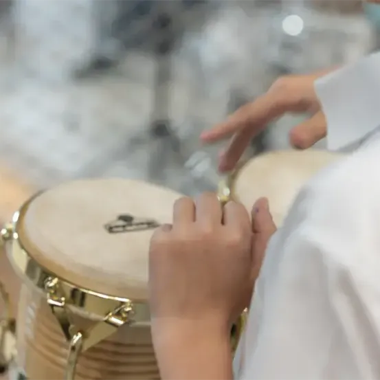 Eine Person spielt eine Trommel mit beiden Händen. Die Trommel hat einen goldenen Rand und eine weiße Oberfläche. Die Person trägt ein weißes Hemd.
