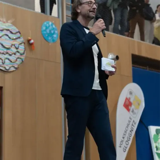 Ein Mann steht auf einer Bühne, hält ein Mikrofon und ein Papier in der Hand, trägt eine Brille und einen Anzug, mit einem Publikum hinter ihm.