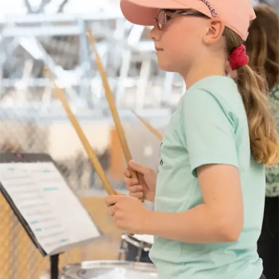 Ein Mädchen übt mit ihren Trommelstöcken, steht neben einem Notenständer mit Musikblättern. Sie trägt ein mintgrünes T-Shirt und einen rosa Hut. Im Hintergrund sieht man verschwommen eine weitere Person und einen Teil eines Schlagzeugs.