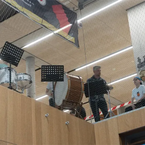 Eine Gruppe von Musikern tritt in einem Innenbereich auf. Eine Person spielt ein Schlagzeug, während eine andere Person auf einer Galerie mit einem Schlagzeug sitzt. Über ihnen hängt ein Banner.