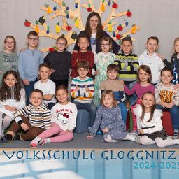 Eine Gruppenaufnahme von Schülern und einer Lehrerin an der Volksschule Gloggnitz, mit einem dekorativen Baum im Hintergrund, für das Schuljahr 2024-2025.