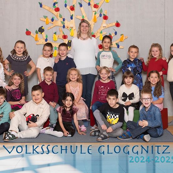 Eine Gruppe von Kindern und eine erwachsene Frau stehen vor einer Wand mit einem Papierbaum und lächeln für ein Foto. Unten im Bild steht 'VOLKSSCHULE GLOGGNITZ 2024-2025.'