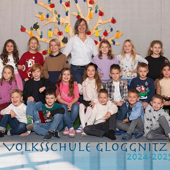 Gruppenfoto von Schülern und einer Lehrerin der Volksschule Gloggnitz, die vor einem geschmückten Baum für das Schuljahr 2024-2025 lächelnd und posierend für die Kamera stehen.