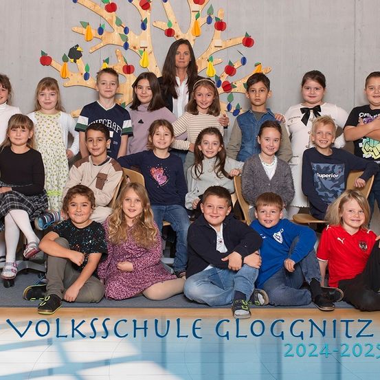 Eine Gruppenaufnahme von Schülern und Lehrern der Volksschule Gloggnitz für das Schuljahr 2024-2025. Die Schüler sind in Reihen angeordnet, lächeln und eine Lehrerin steht in der Mitte.