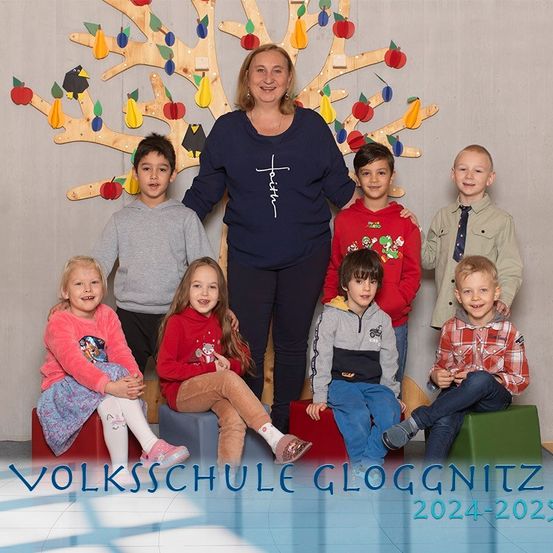 Eine Gruppe von Kindern und einer erwachsenen Frau posiert für ein Foto vor einem dekorativen Baum, wahrscheinlich an der Volksschule Gloggnitz, für das Schuljahr 2024-2025.