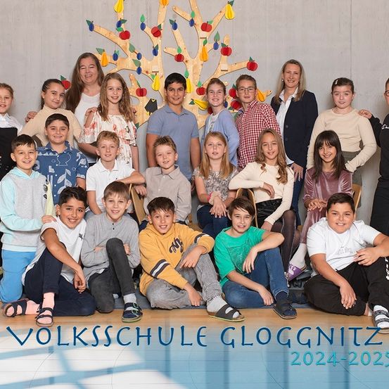 Eine Gruppe von Schülern und Lehrern posiert für ein Foto an der Volksschule Gloggnitz, mit einem Baumdesign im Hintergrund und den Jahren 2024-2025 am unteren Rand angezeigt.