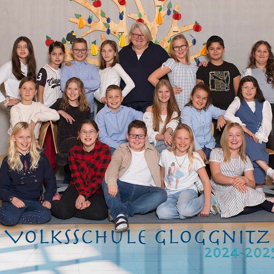 Eine Gruppe von Kindern und einer erwachsenen Frau posiert für ein Foto vor einem Baum mit Äpfeln. Unten stehen die Worte Volksschule Gloggnitz 2024-2025 geschrieben.