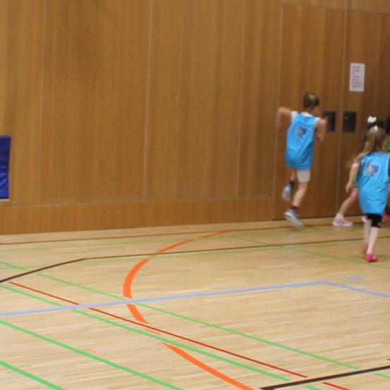 Ein verschwommenes Bild zeigt eine Gruppe junger Mädchen, die auf einem Holzboden mit bunten Linien laufen. Ein Mädchen vorne trägt ein blaues Trikot.