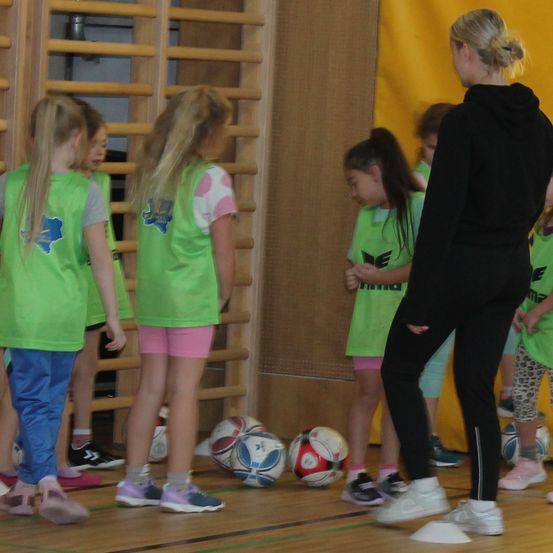 Eine Gruppe von Kindern in grünen Westen steht auf einem Holzboden in einer Turnhalle. Sie betrachten Fußballbälle auf dem Boden. Eine Frau in Schwarz steht vor ihnen.